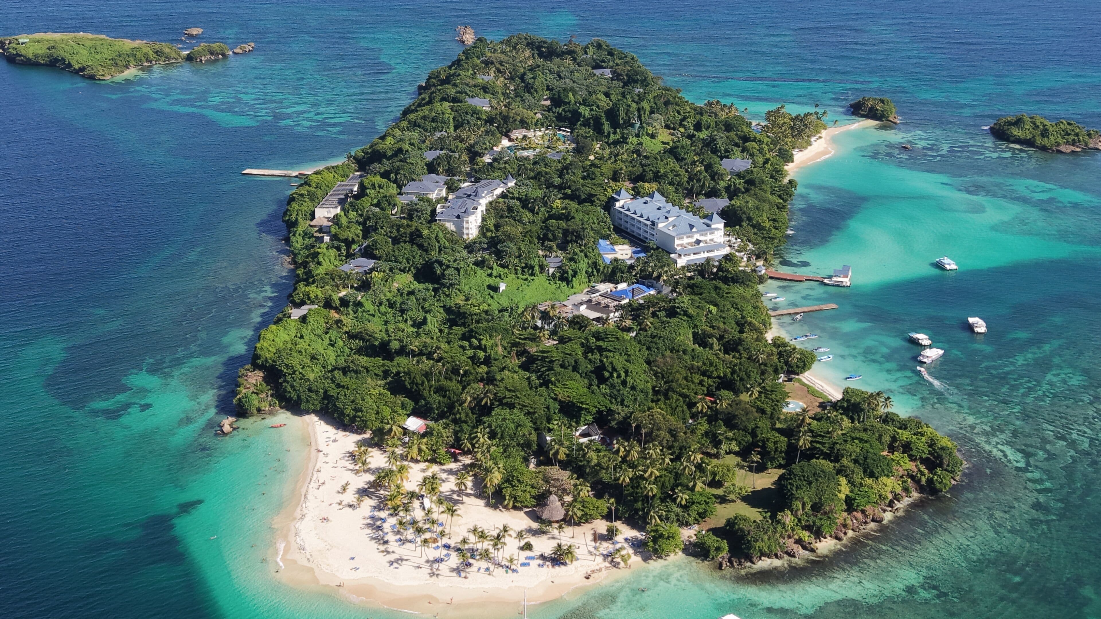 An island with a beach, clear turquoise waters, and a resort with multiple buildings.