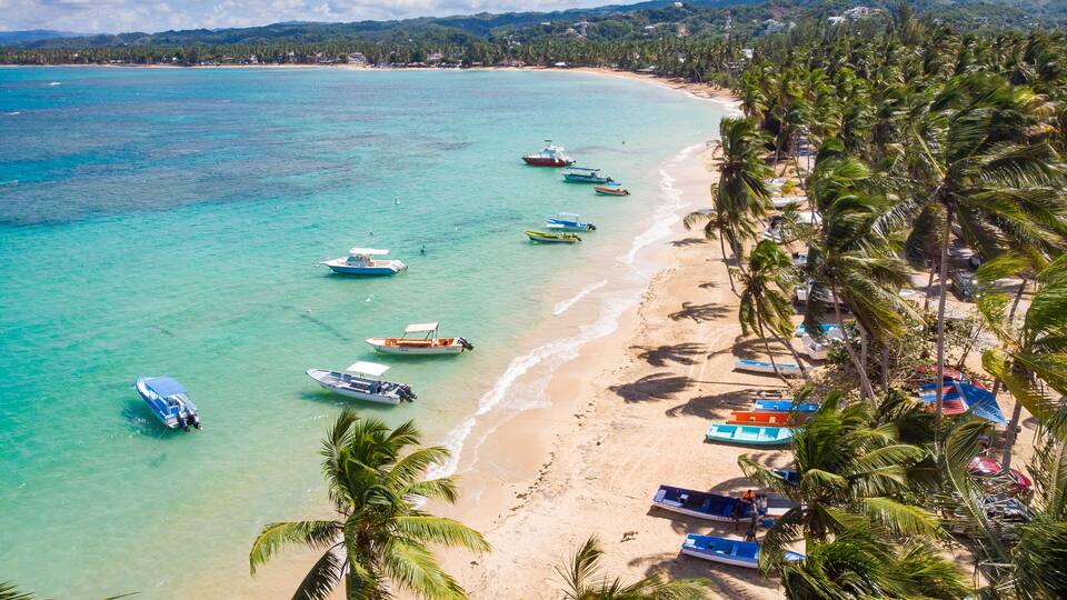 A tropical beach with clear turquoise waters, several boats anchored near the shore, and a line of palm trees along the coastline.