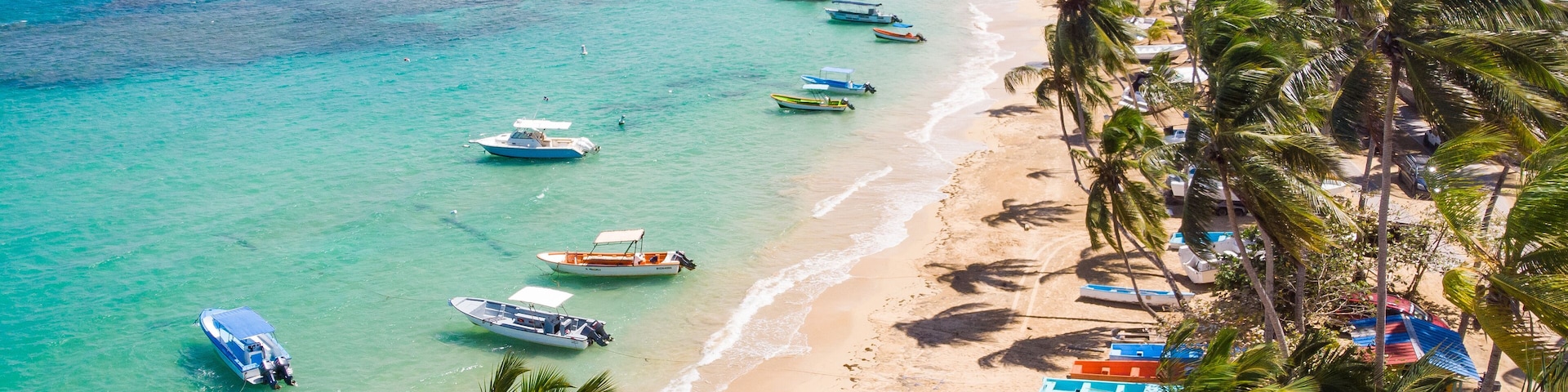 A tropical beach with clear turquoise waters, several boats anchored near the shore, and a line of palm trees along the coastline.