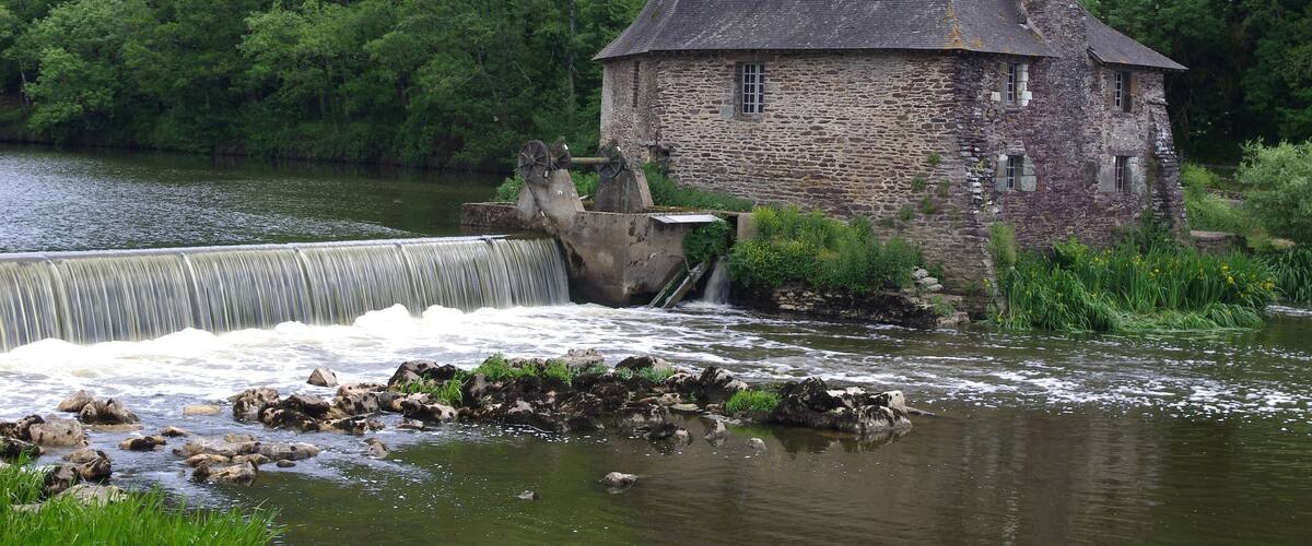A stone house with a water wheel by a river, surrounded by lush greenery and a rocky hill.