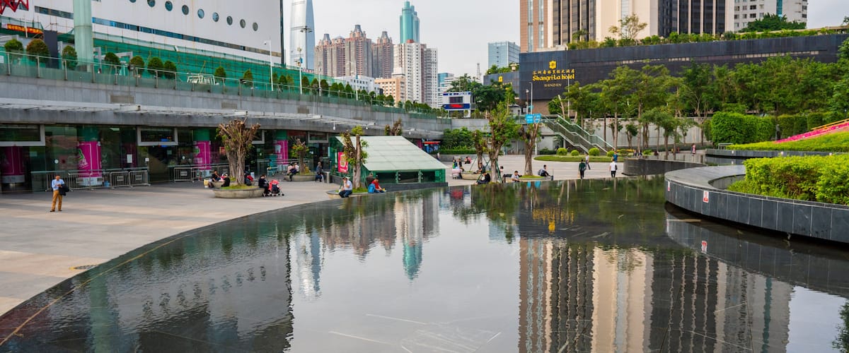 A city square with a reflective water feature, modern buildings, and a covered seating area.