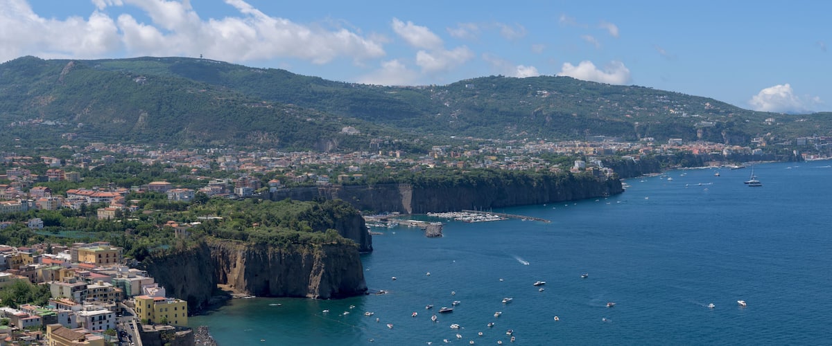 A coastal city with a prominent cliff, a harbor with boats, and a mountainous backdrop.