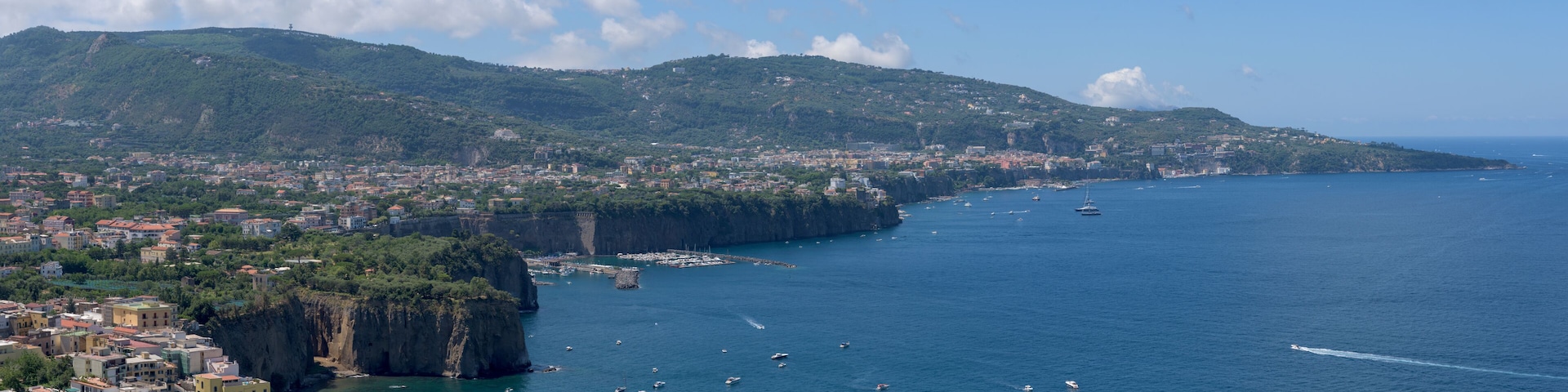 A coastal city with a prominent cliff, a harbor with boats, and a mountainous backdrop.