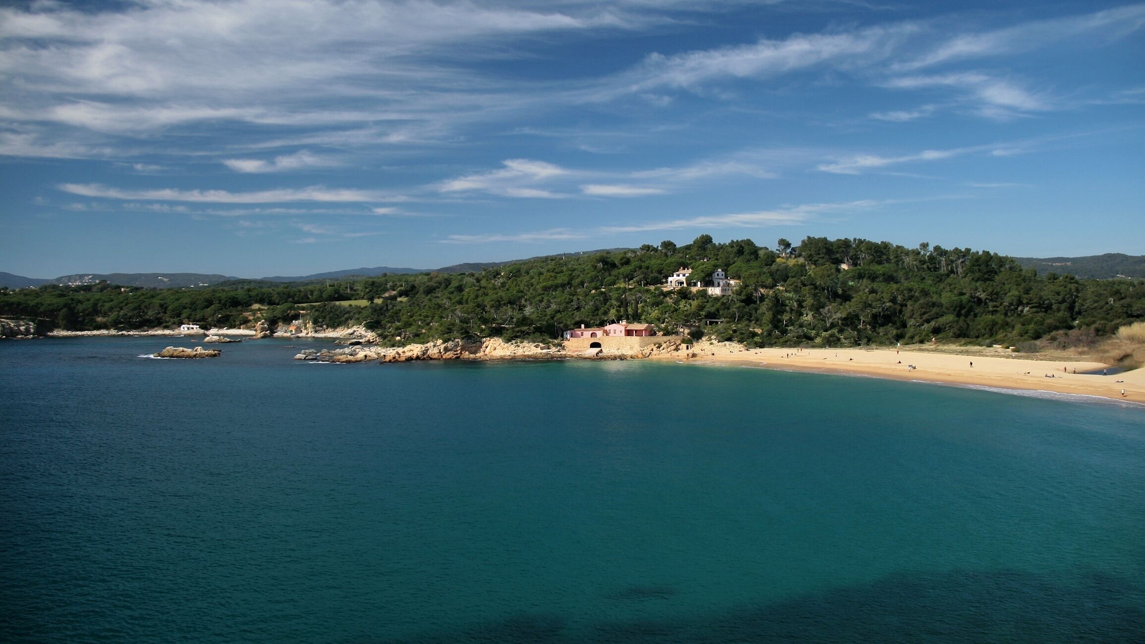 A coastal landscape with a sandy beach, clear turquoise waters, and a forested hill in the background.