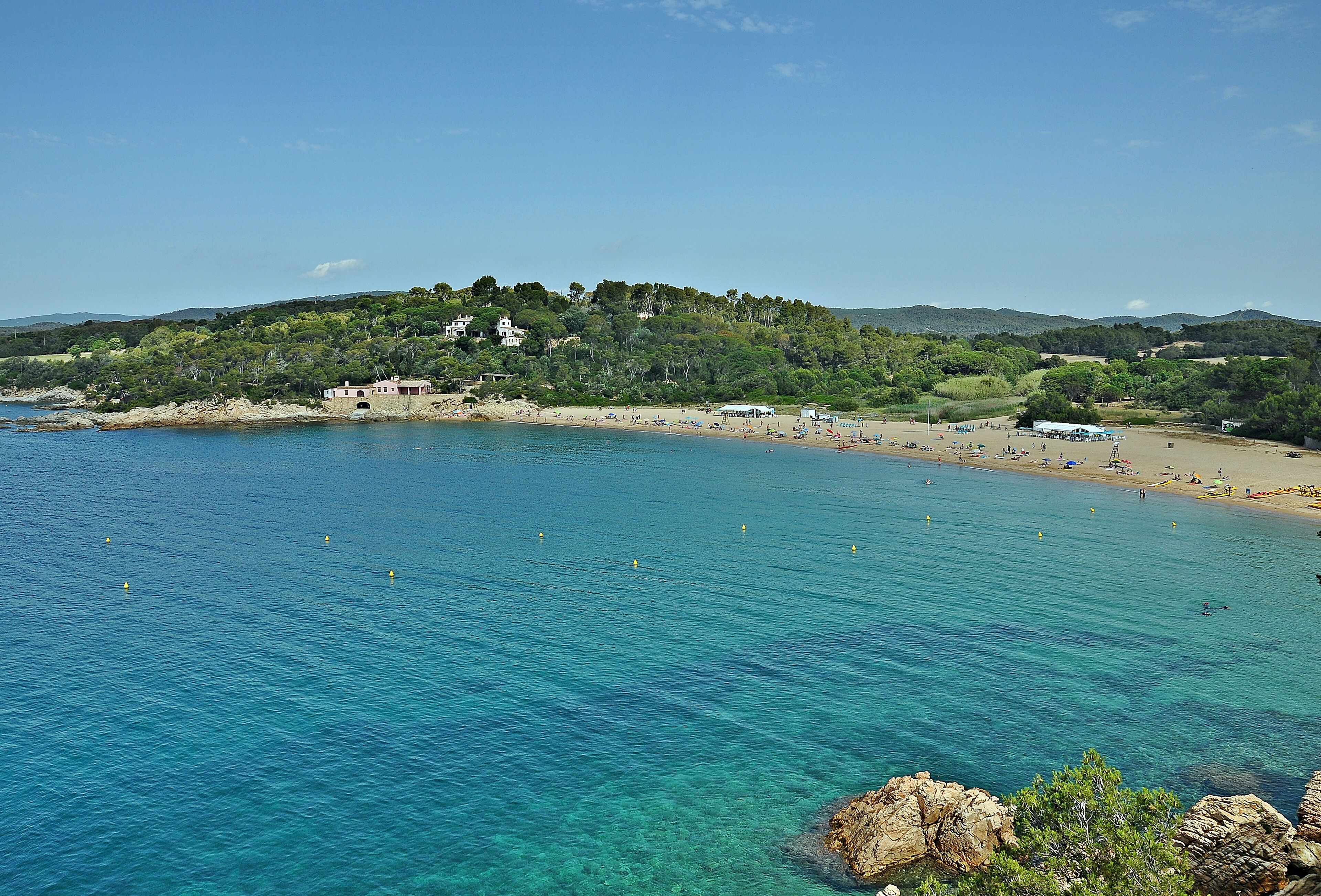 A coastal landscape with clear blue waters, a sandy beach, and a hill with buildings and greenery.