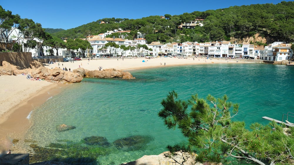 A coastal town with clear turquoise waters, sandy beach, and buildings on a hillside.