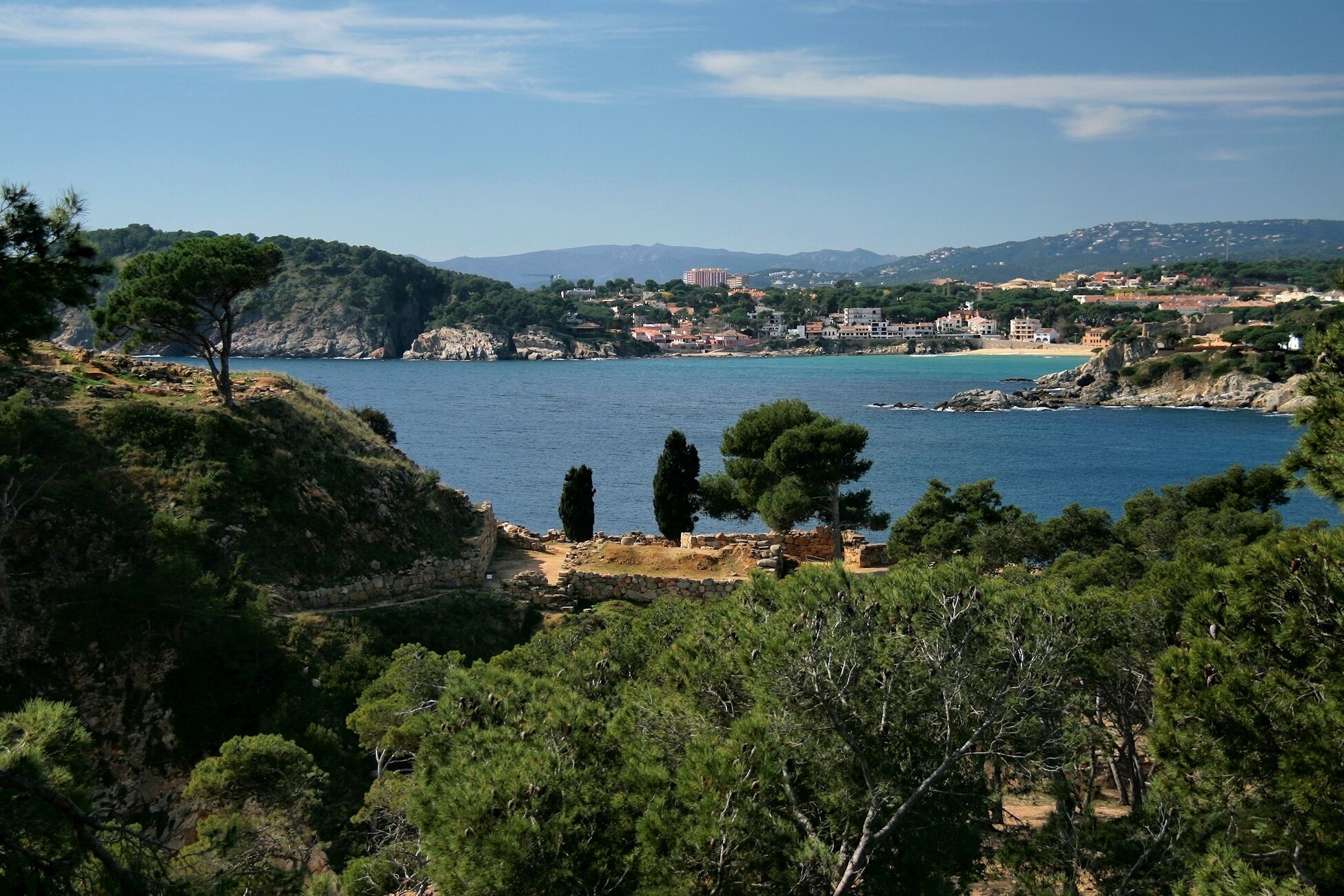 A coastal landscape with a clear blue sea, rocky cliffs, and a small village with buildings in the background.