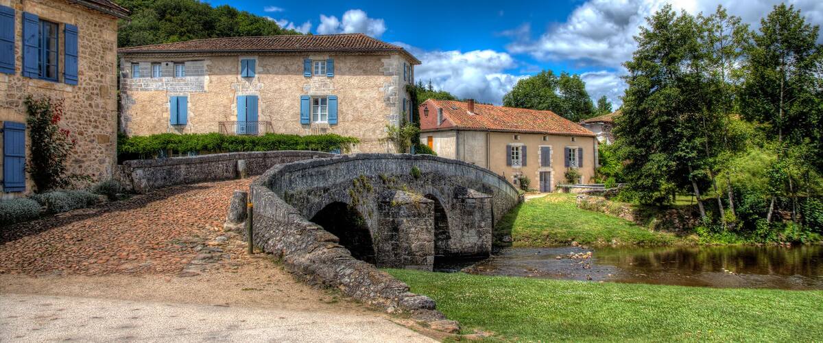A stone bridge over a stream, surrounded by stone buildings with blue shutters, set against a backdrop of lush green hills and a partly cloudy sky.
