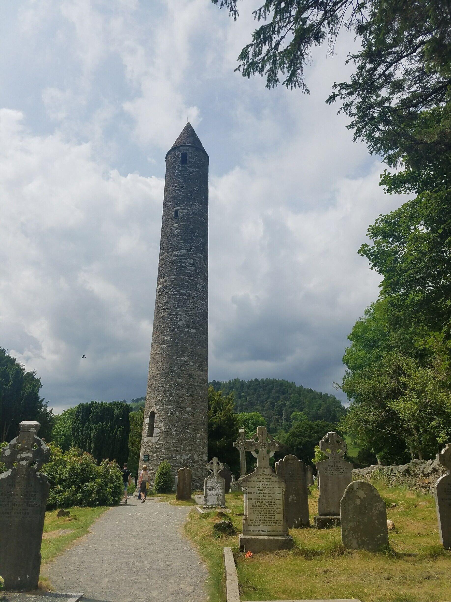Une haute tour de pierre se dresse dans un cimetière, au milieu des pierres tombales, et un chemin y mène.