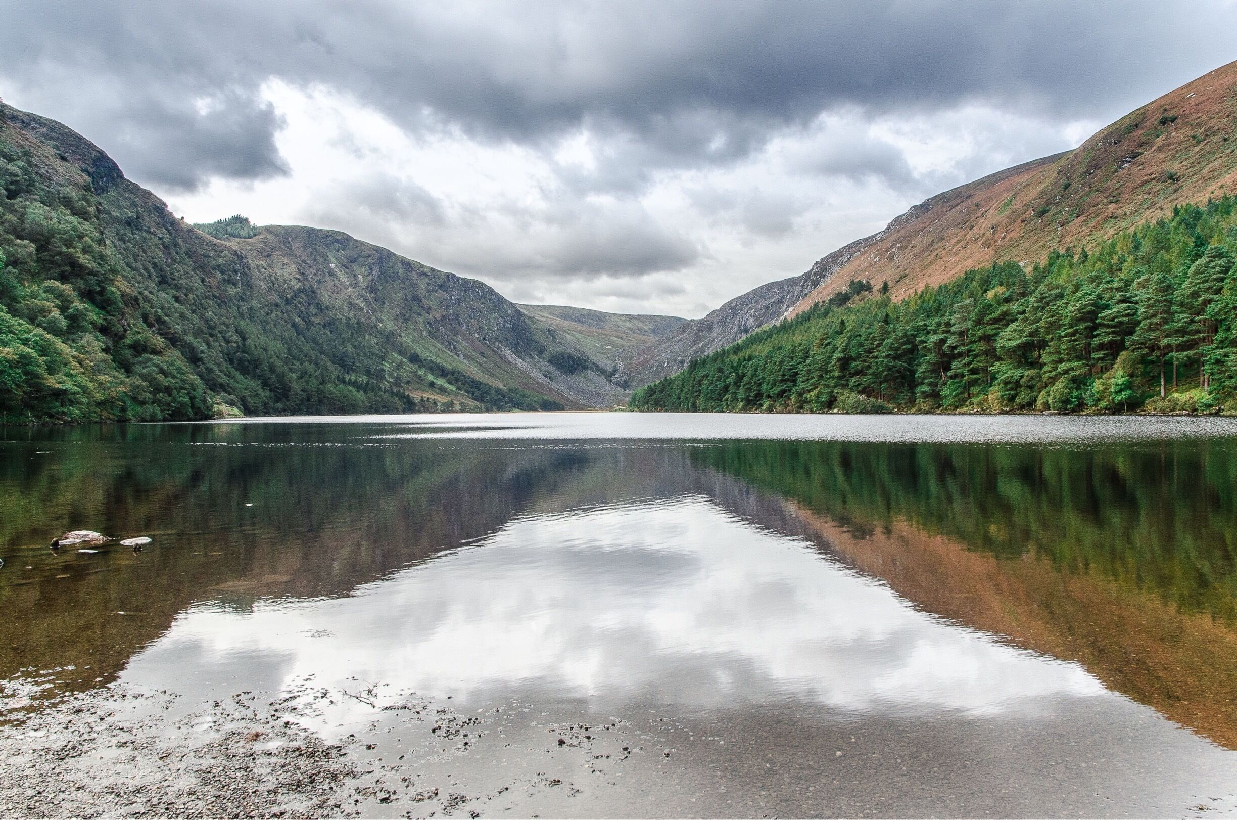 A calm lake surrounded by forested hills under a cloudy sky.