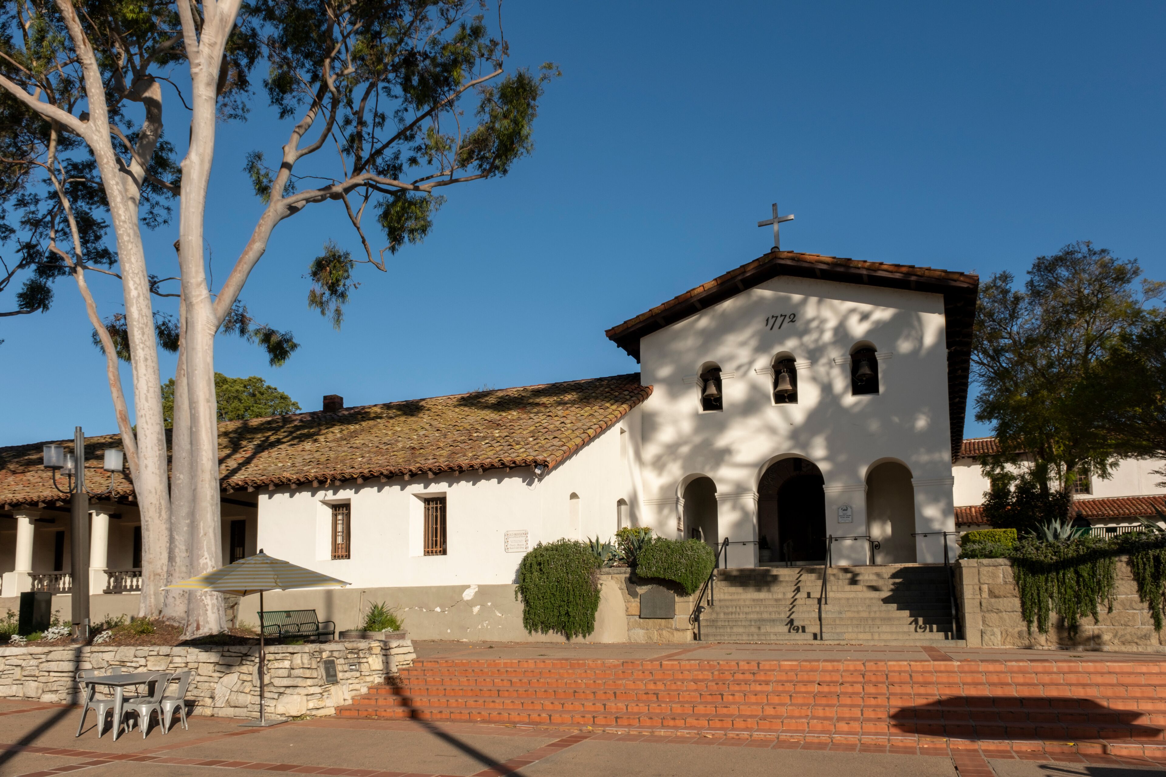 A white church with a cross on top, a stone bench, and a tree in front.