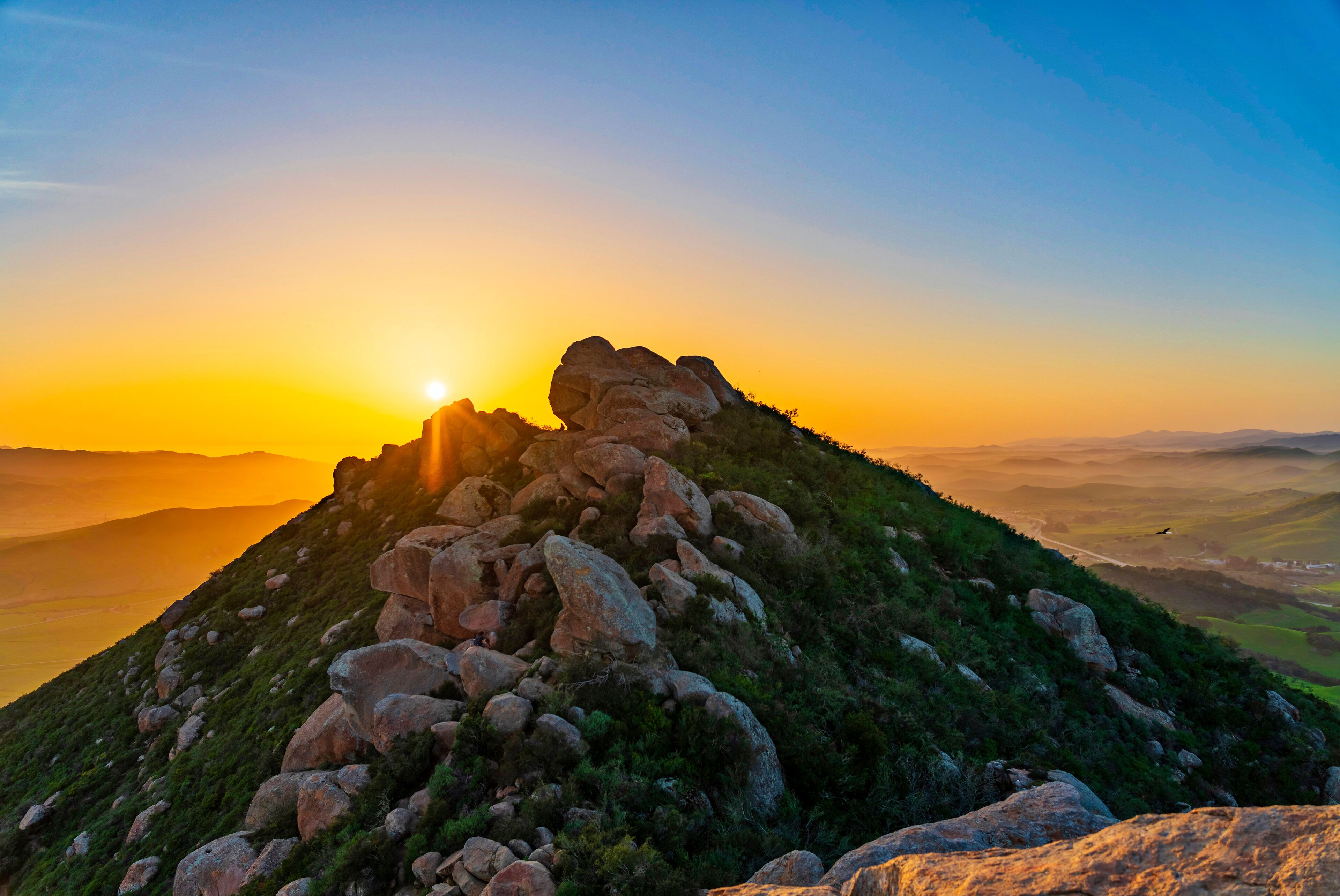 A rocky mountain peak at sunset with the sun setting behind it.