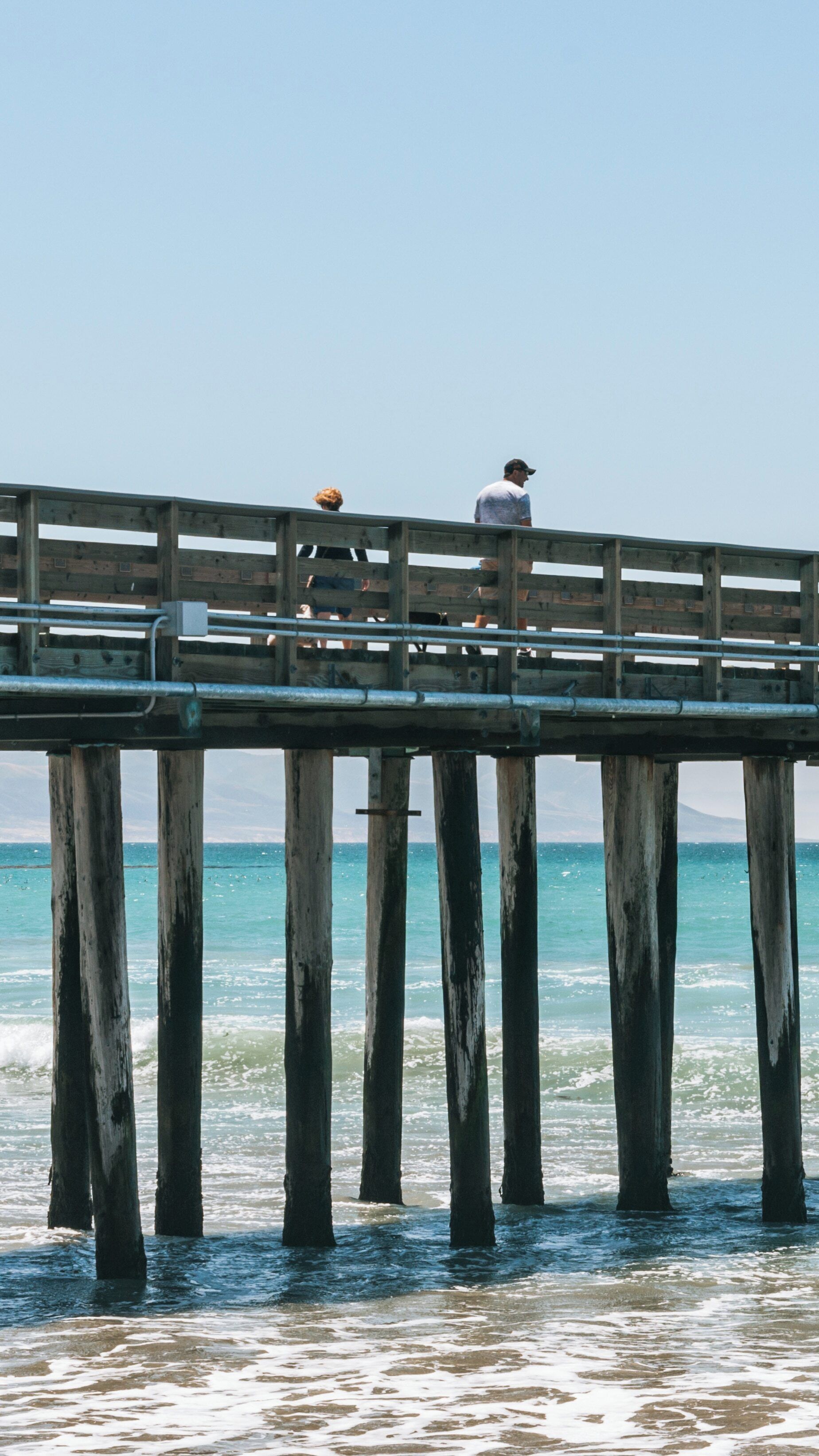 Two people walking on a wooden pier at Cayucos State Beach in California on a sunny day with clear blue skies and gentle ocean waves