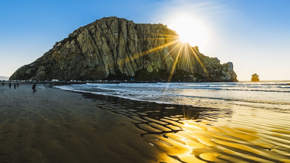 A rocky cliff by the sea with the sun shining on the sand.