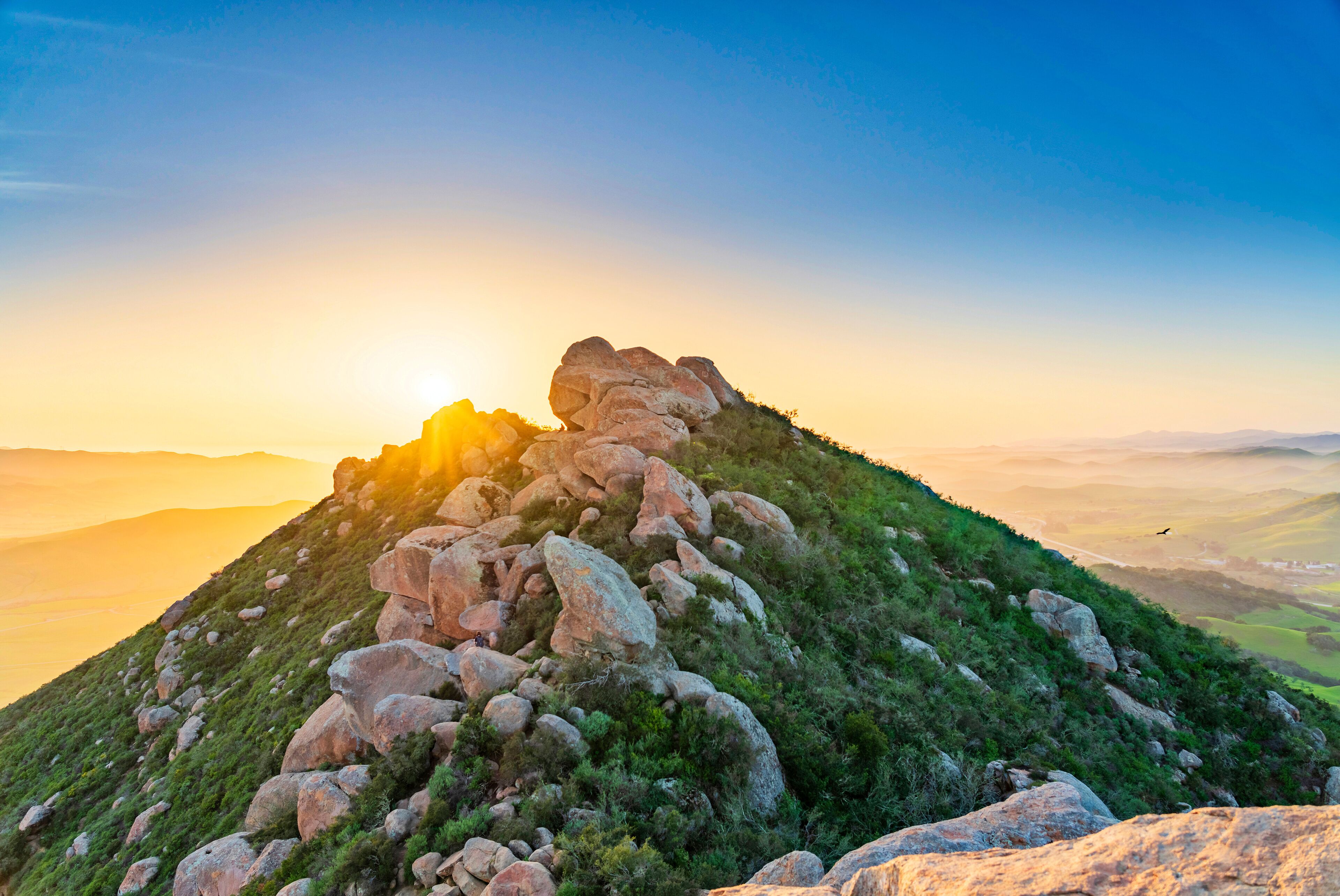 A rocky hill with a prominent rock formation at the peak, surrounded by greenery and a clear sky.