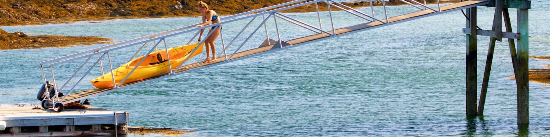 A wooden bridge over water with a yellow kayak, boats, and a motel in the background.