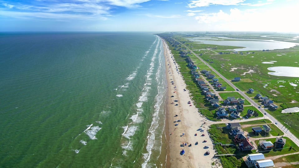 An aerial view of a long, narrow beach with a row of houses and a road parallel to the shore.
