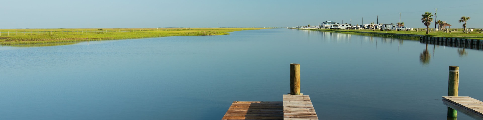 A wooden pier extends into a calm body of water under a clear blue sky at the Blue Water RV Beach Resort, Surfside Beach, Texas. The tranquil water seems to merge with the blue summer sky.