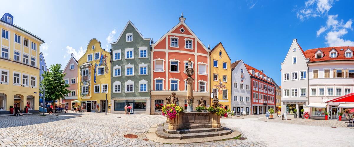 A cobblestone square with colorful historic buildings, a central fountain, and a clear blue sky.