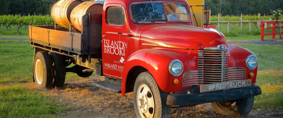 A vintage red truck with barrels on the back, parked on a dirt road in a rural area with trees and a wooden fence in the background.
