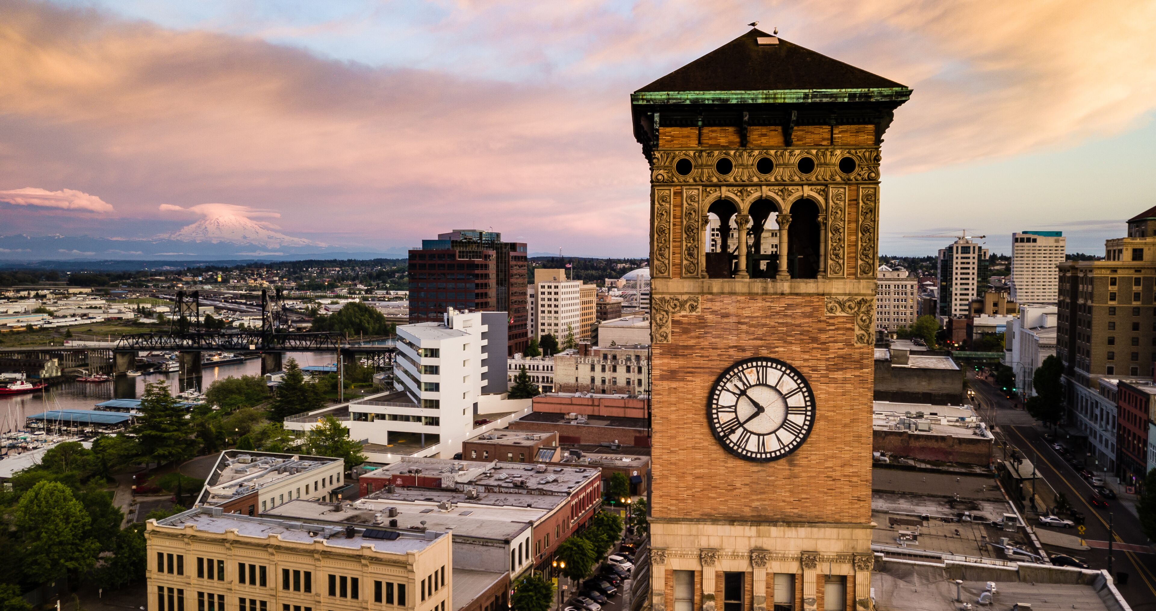 A clock tower with a large clock face, surrounded by urban buildings and a bridge.