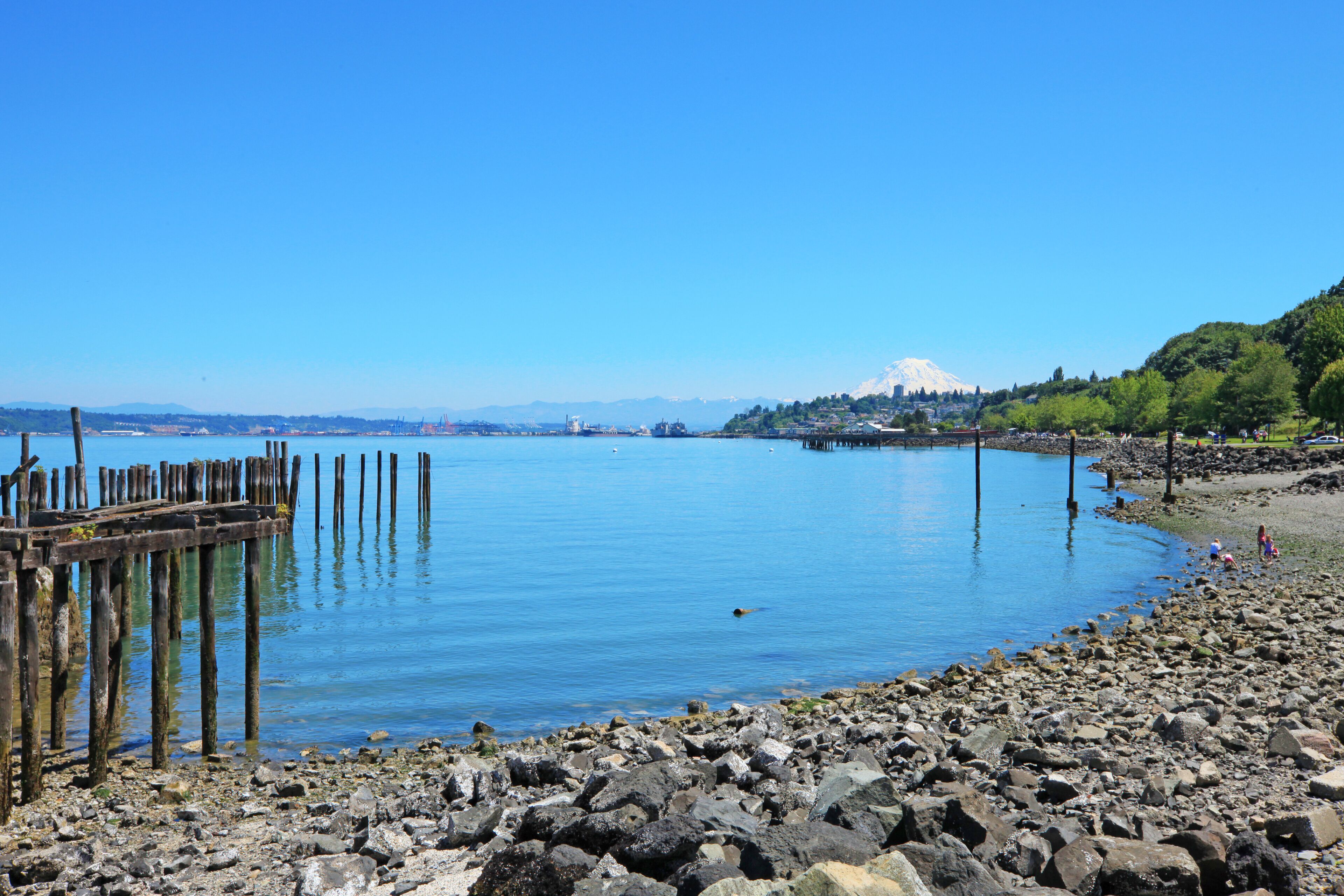 A rocky shoreline with a wooden pier extending into calm blue water. In the background, a mountain with a snow-capped peak is visible.