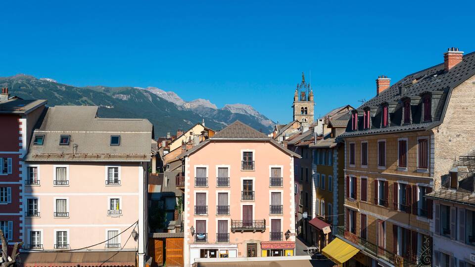 A street with colorful buildings, a church tower, and mountains in the background.