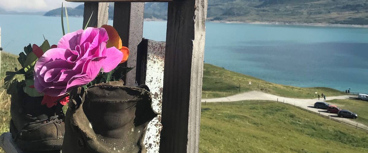 A wooden sign with 'Refuge du Petit Ménis' written on it, a pair of boots with a pink flower, and a scenic view of a lake and mountains in the background.