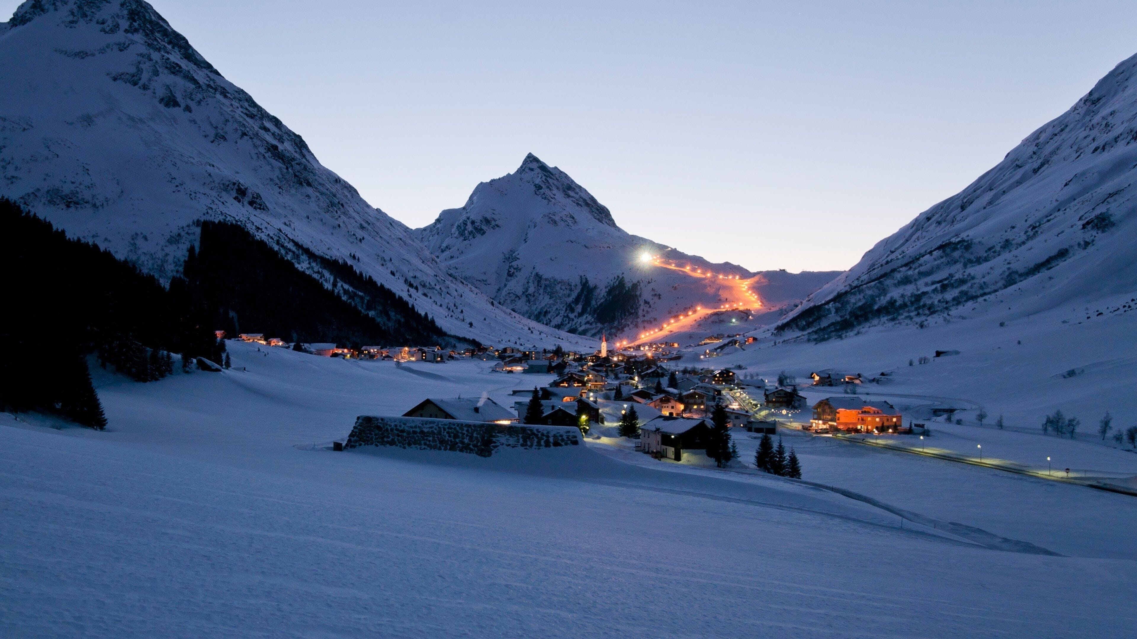 A snow-covered mountain village at dusk with illuminated buildings and a church spire.