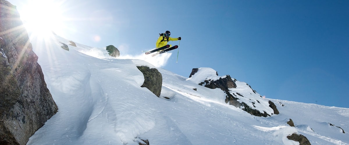 A skier in mid-air performing a jump over a snowy mountain with rocks.