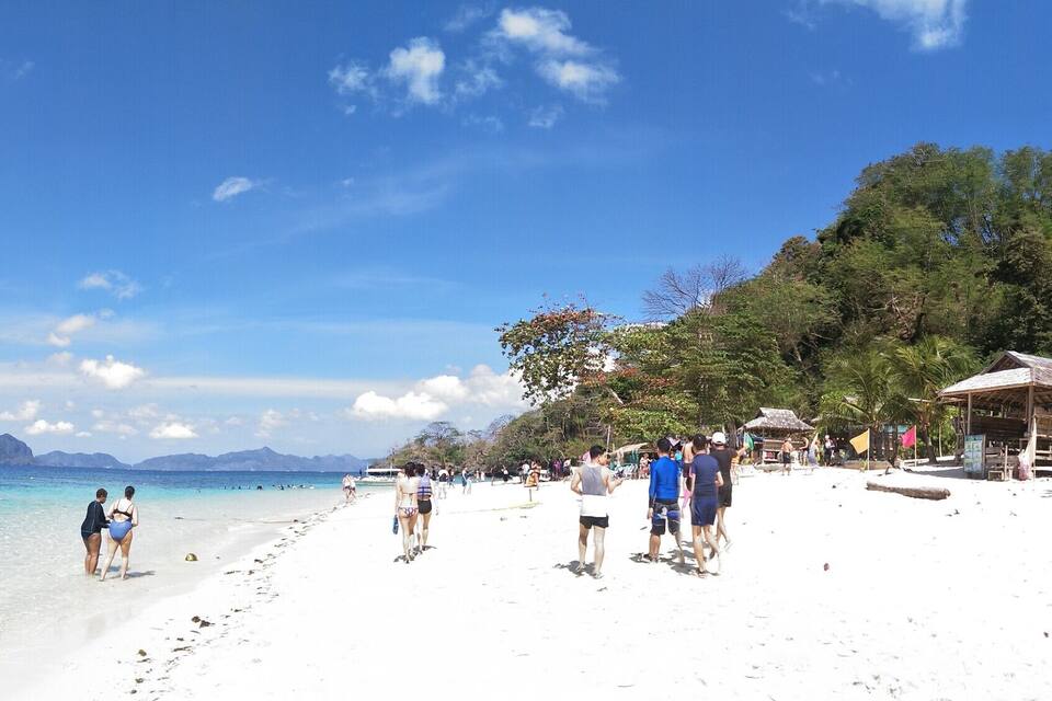 Une plage bordée d’une eau turquoise et limpide, un bateau amarré sur le rivage et une rangée de cabanes le long de la plage.