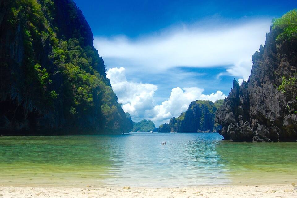 A coastal landscape with clear turquoise waters, a sandy beach, and lush green cliffs.