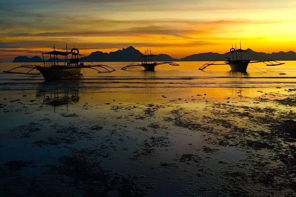 Un atardecer sobre una superficie de agua tranquila, con tres barcos anclados en primer plano.