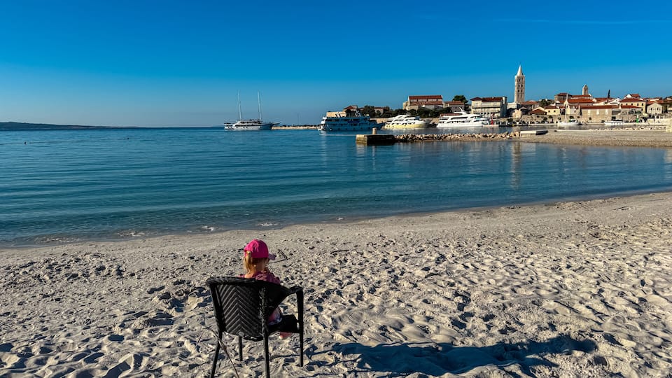A sandy beach with a person sitting on a chair, a clear blue sky, and a harbor with boats and buildings in the background.