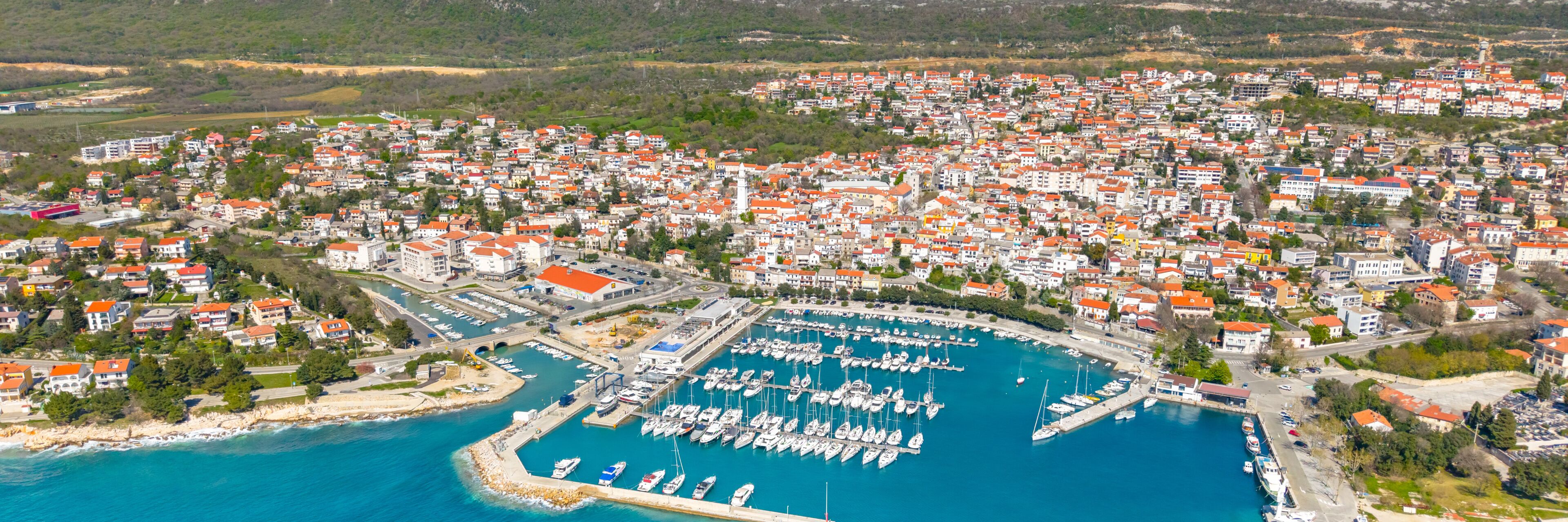 An aerial view of a coastal town with a marina filled with boats, surrounded by residential buildings with red roofs.