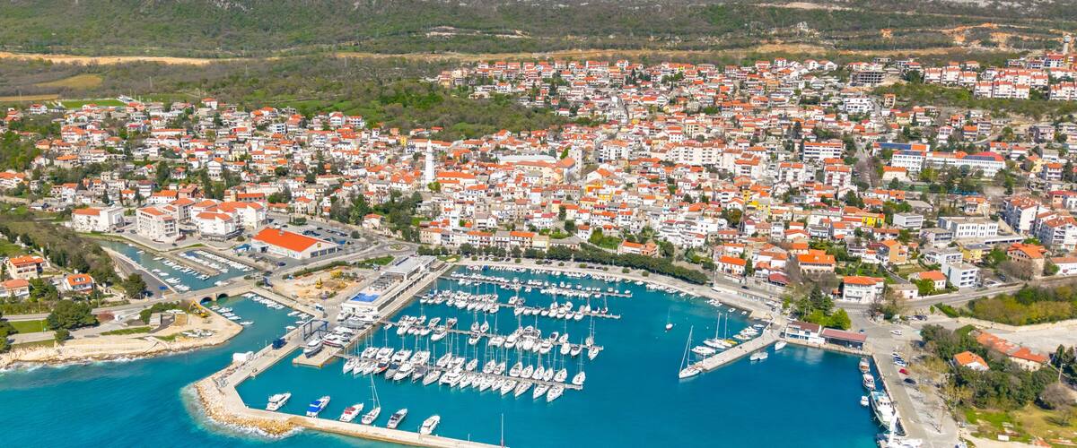 An aerial view of a coastal town with a marina filled with boats, surrounded by residential buildings with red roofs.