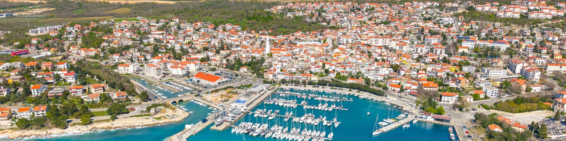 An aerial view of a coastal town with a marina filled with boats, surrounded by residential buildings with red roofs.