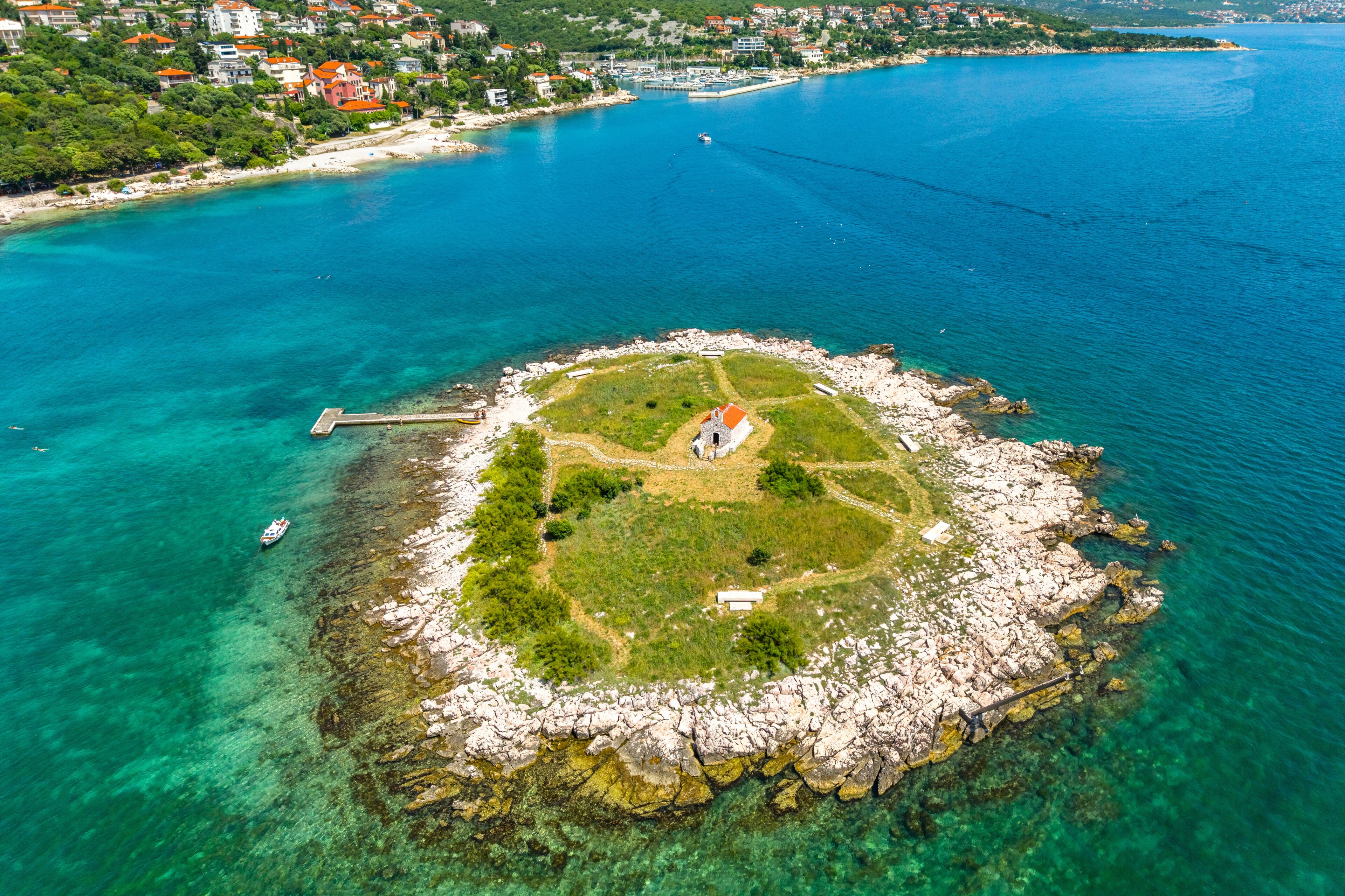 Drone image of a small rocky island San Marino near Novi Vinodolski, featuring a red-roofed chapel in the center, surrounded by green grass and crystal-clear turquoise waters