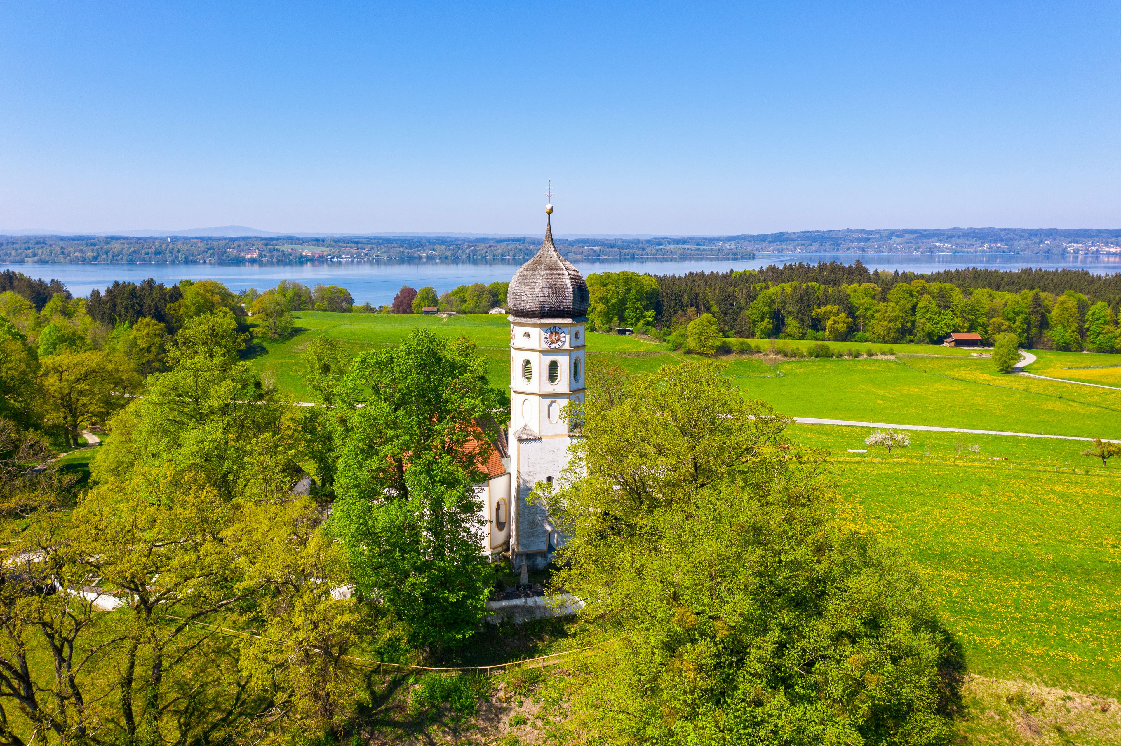 Een witte kerk met een koepel en klokkentoren, omringd door weelderige groene velden en een meer.