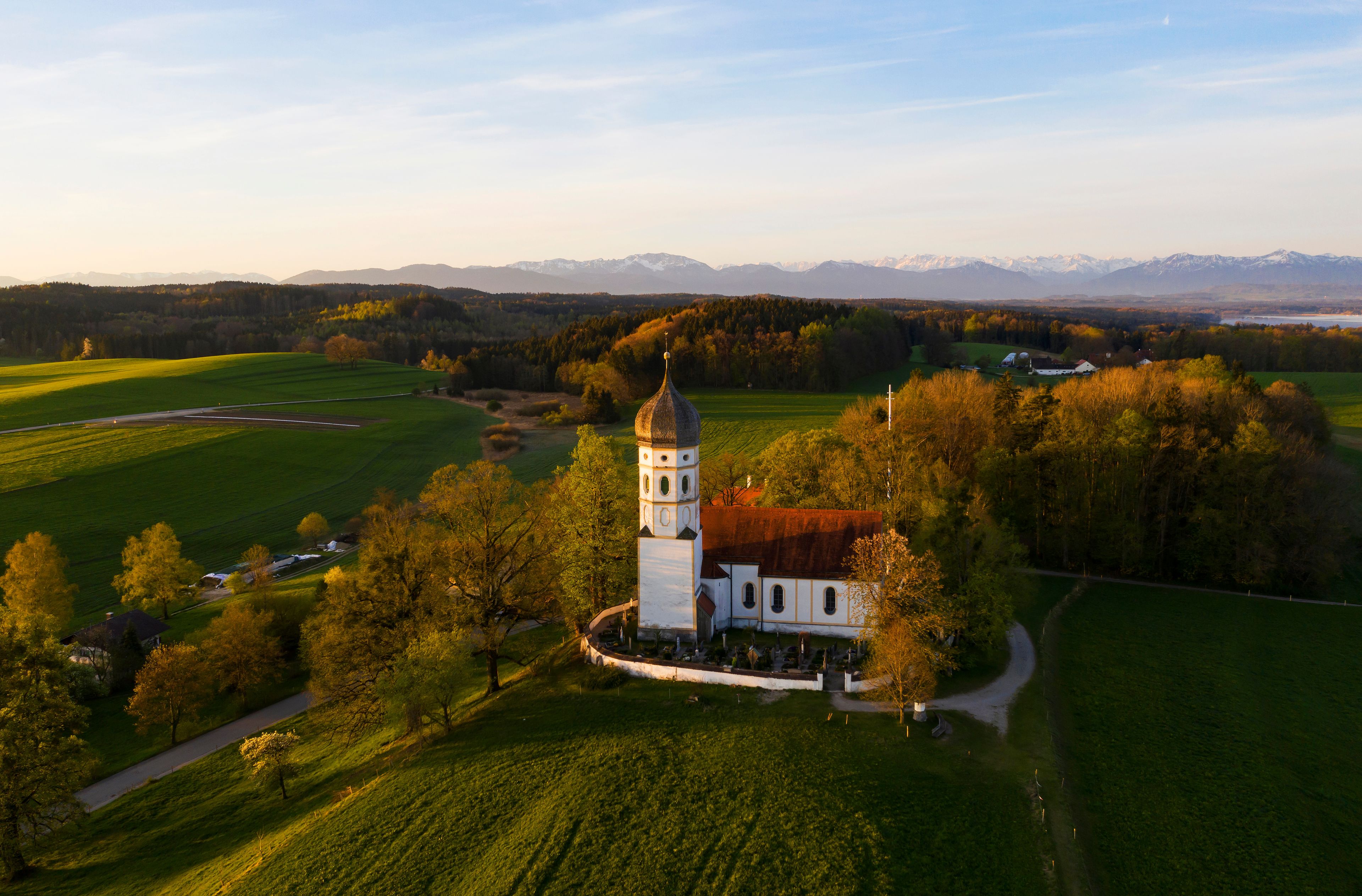 Een kerk met een kerktoren, omringd door groene velden en bomen, met bergen op de achtergrond.