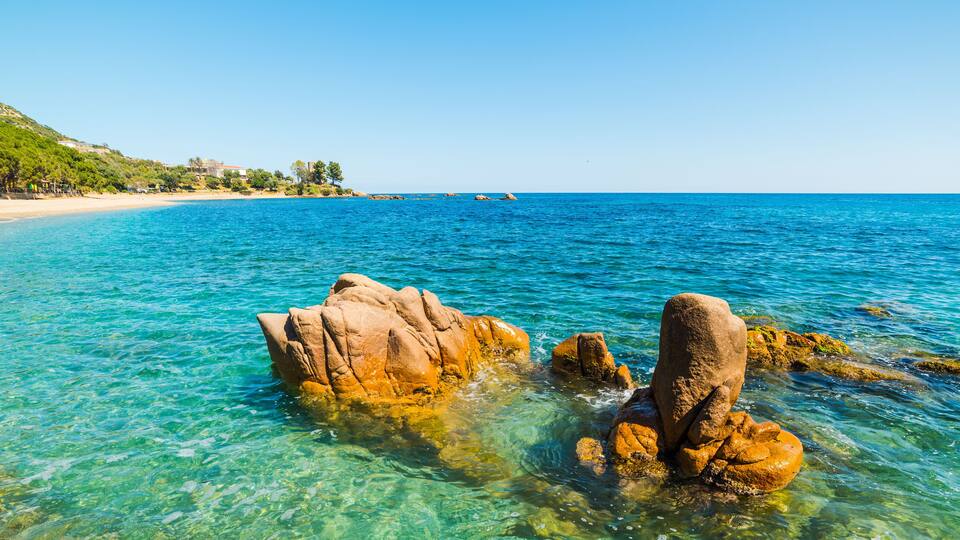 A clear blue sea with rocky formations in the foreground and a sandy beach with trees in the background.