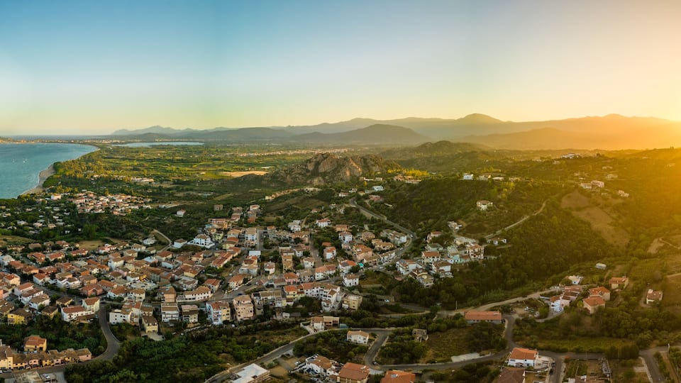 A coastal town with a dense cluster of houses, a coastline, and distant mountains under a sunset sky.