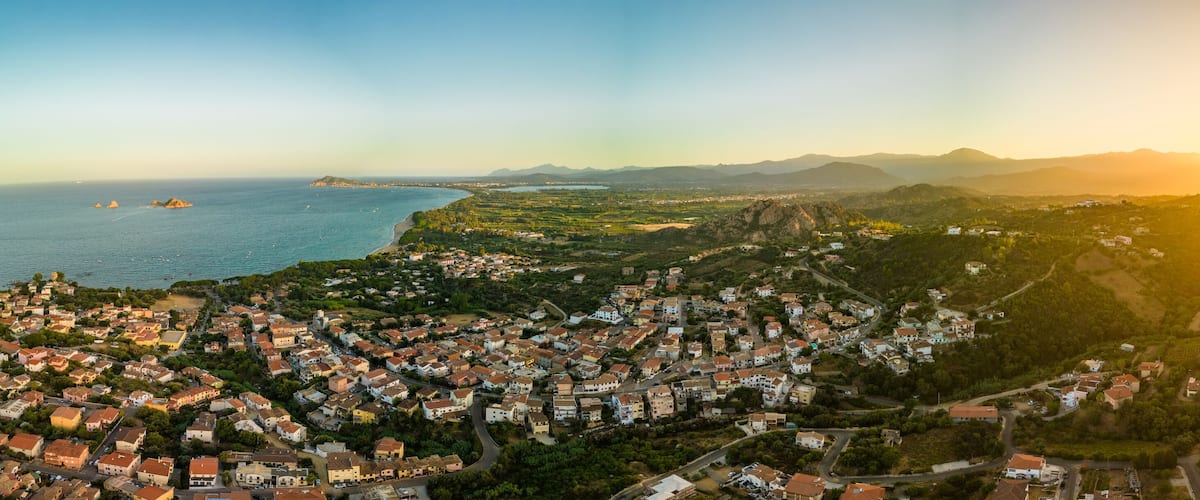 A coastal town with a dense cluster of houses, a coastline, and distant mountains under a sunset sky.