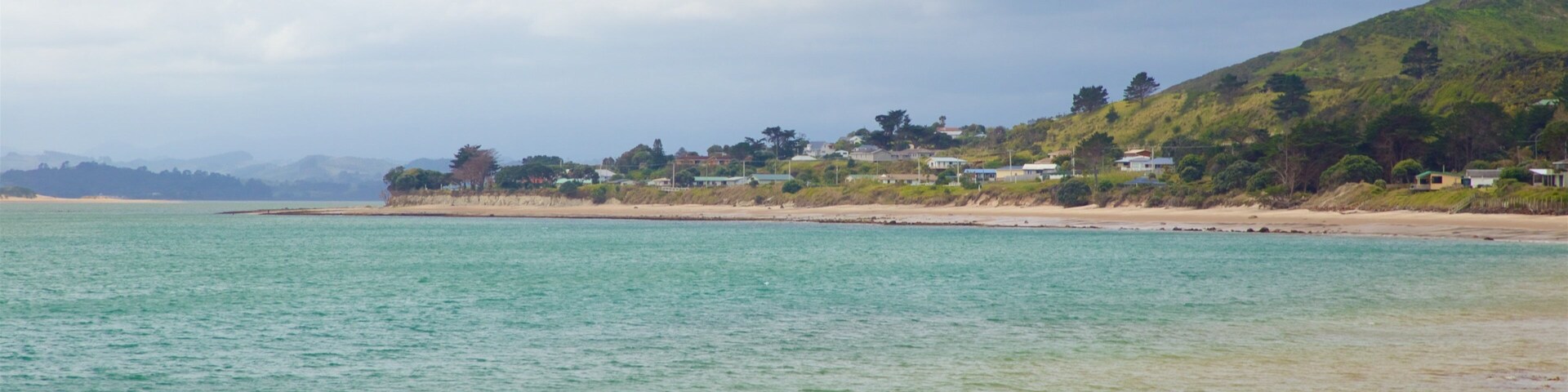 Paisaje costero con aguas turquesas cristalinas, playa arenosa y una colina con casas.