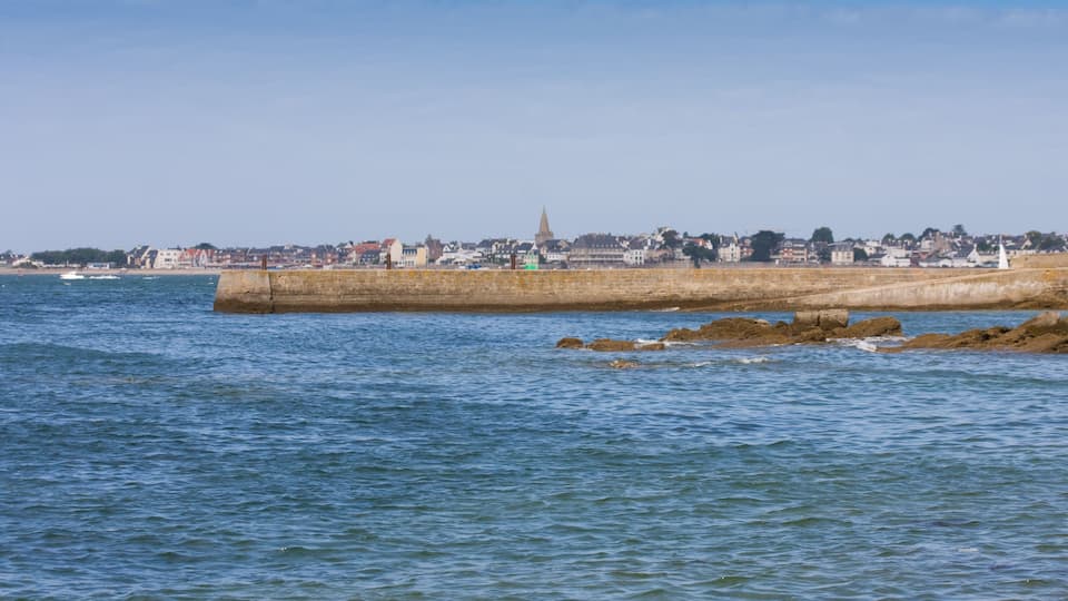 A coastal cityscape with a prominent church spire, a stone breakwater, and a calm sea.
