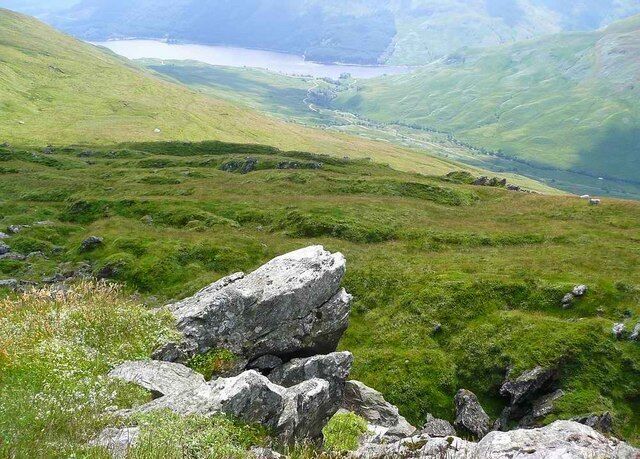 A mountainous landscape with a lake in the distance, rocky outcrops, and lush green vegetation.