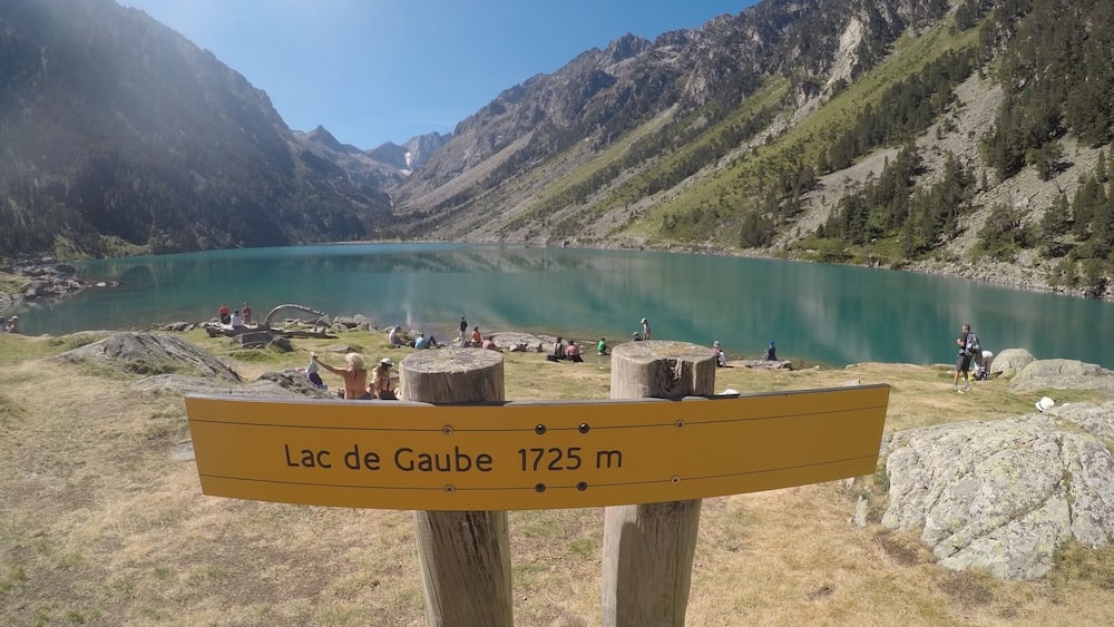 Un panneau indiquant le lac de Gaube, situé à 1 725 mètres d’altitude, entouré de montagnes et bordé d’un lac aux eaux turquoise, avec des personnes présentes aux alentours.