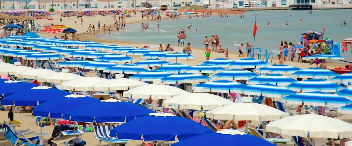 A crowded beach with numerous blue and white umbrellas, people sunbathing, and a clear blue sky.