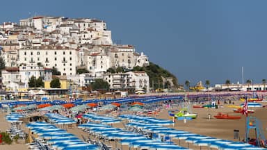 A crowded beach with numerous blue and white umbrellas, a sandy shore, and a backdrop of white buildings on a hillside.