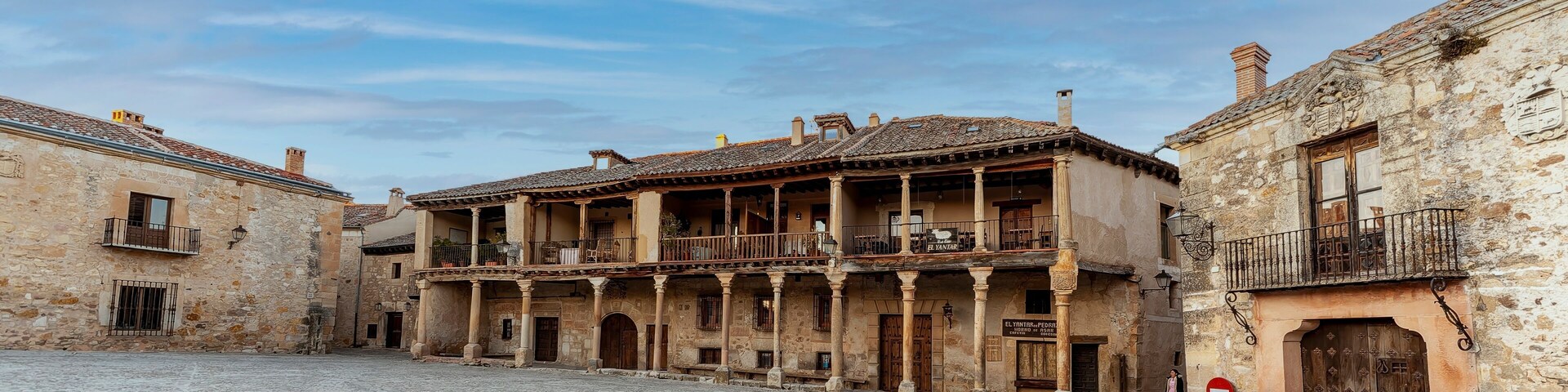 Uma rua de paralelepípedos com edifícios históricos de pedra, incluindo um edifício com varanda e uma placa com a inscrição